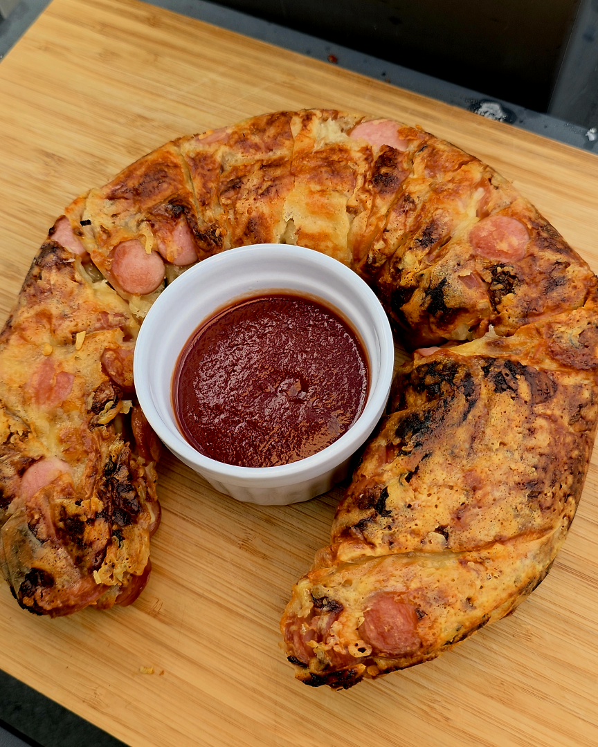 Goldbraun gebackene, ringförmige Hot Dogs im Donut-Style, umwickelt mit Blätterteig und garniert mit Ketchup und Senf in der runden Omnia Backform.