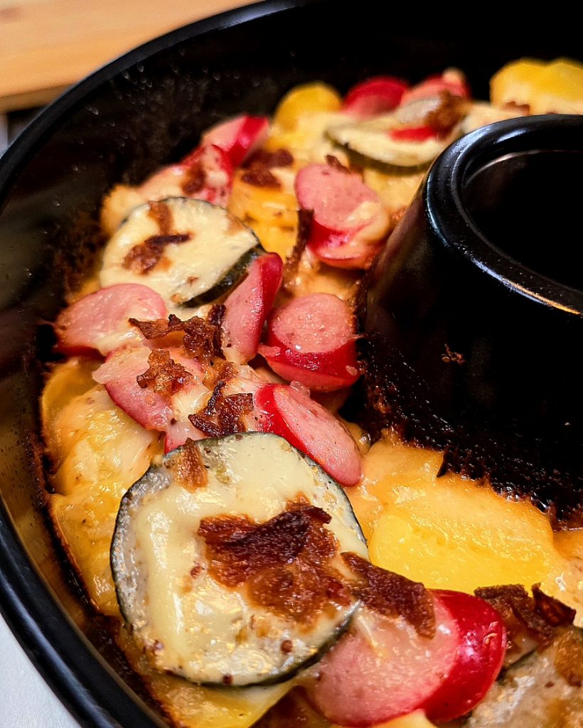 Draufsicht auf einen fertig gebackenen Kartoffelauflauf im runden Omnia-Campingbackofen. Man sieht goldgelbe Kartoffelscheiben, Hot-Dog-Wursträdchen, geschmolzenen Käse und Röstzwiebeln