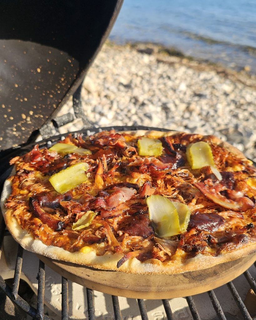 Pizza Pulled Pork Big green egg Minimax Grill Eine fertig gebackene Pizza mit Pulled Pork, Käse, roten Zwiebeln und Barbecue-Soße auf einem Pizzastein, zubereitet auf einem Big Green Egg MiniMax Grill.